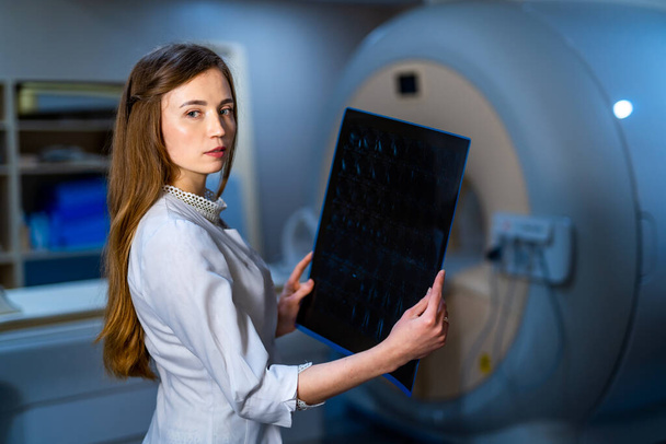 Young seriuos competent female doctor looking at X-ray at doctor's office. Computer tomography background. - Φωτογραφία, εικόνα