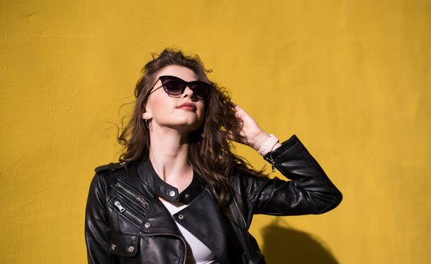 pretty brunette smiling girl in leather jacket in sunglasses on bright orange background. young happy woman on sunny day in city - Photo, Image