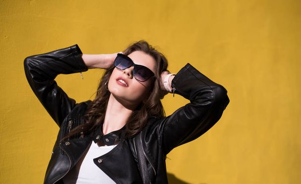pretty brunette smiling girl in leather jacket in sunglasses on bright orange background. young happy woman on sunny day in city - Фото, изображение