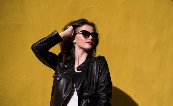 pretty brunette smiling girl in leather jacket in sunglasses on bright orange background. young happy woman on sunny day in city - Photo, image