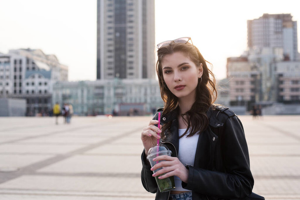 young cheerful brunette student girl is enjoying with drink on city background - Photo, Image