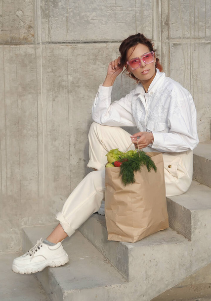 Κομψή όμορφη γυναίκα κρατά τσάντα χαρτί χειροτεχνίας με λαχανικά που θέτουν σε εξωτερικούς χώρους. - Φωτογραφία, εικόνα