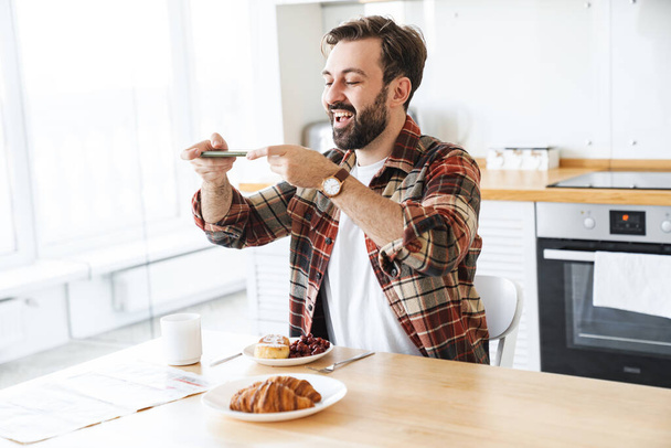 Portrait of joyful bearded man smiling and taking photo his breakfast on cellphone in kitchen at home - Foto, Bild