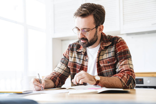 Porträt eines jungen bärtigen Mannes mit Brille, der zu Hause mit Notizen arbeitet - Foto, Bild