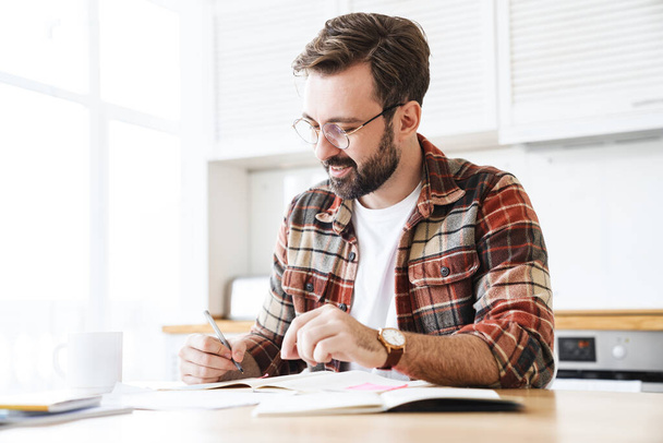 Portrait of excited bearded man wearing eyeglasses smiling while working with notes at home - Фото, изображение