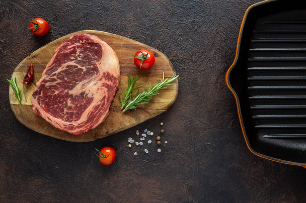 Raw meat Black Angus for Rib eye or Striploin steak on wooden cutting board with fresh rosemary,cherry tomatoes and dried papper.And half of cast iron grill pan.Top view. - Φωτογραφία, εικόνα