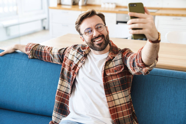 Portrait of young happy bearded man smiling and using cellphone while sitting on sofa at home - Foto, Bild
