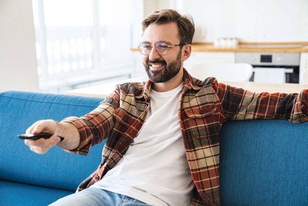 Portrait of young happy bearded man smiling and watching television while sitting on sofa at home - Photo, image