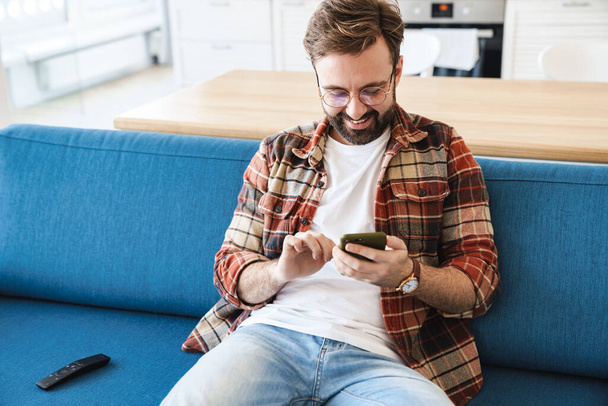 Portrait of young happy bearded man smiling and using cellphone while sitting on sofa at home - Photo, image