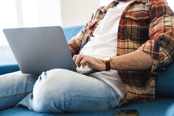 Portrait of young caucasian man wearing plaid shirt using laptop while sitting on sofa at home - Foto, immagini