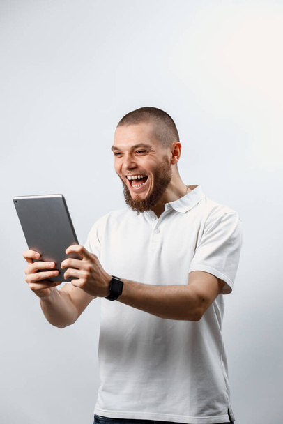 Retrato de un guapo barbudo con una camiseta blanca hablando de una tableta de video llamada. aislado. emociones
 - Foto, imagen