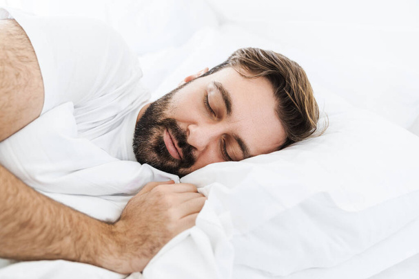 Image of young unshaven caucasian man sleeping alone in bed with white linen at home - Foto, afbeelding