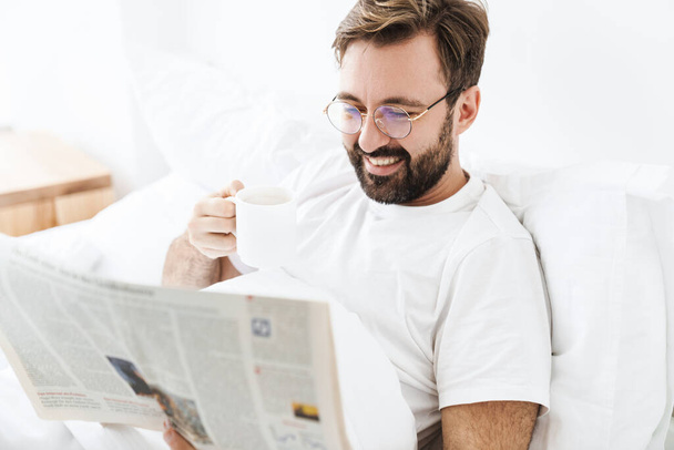 Image of young unshaven caucasian man drinking coffee and reading newspaper while lying in bed at home - Фото, изображение
