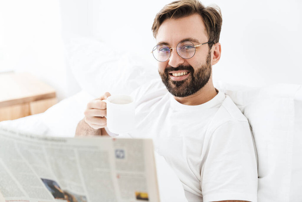 Image of young unshaven caucasian man drinking coffee and reading newspaper while lying in bed at home - Фото, зображення