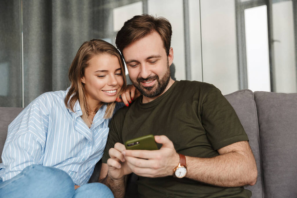 Photo of cheerful beautiful couple smiling and using mobile phone while sitting on sofa at home - Valokuva, kuva