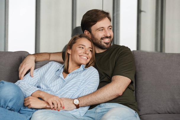 Photo of cheerful caucasian couple smiling and hugging while sitting on sofa at home - Photo, Image