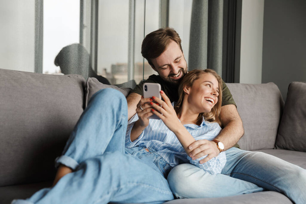 Photo of funny beautiful nice couple laughing and using mobile phone while sitting on sofa at home - Valokuva, kuva