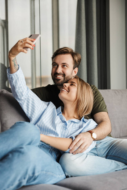 Photo of joyful beautiful nice couple smiling and taking selfie photo on cellphone while sitting on sofa at home - Foto, Imagem
