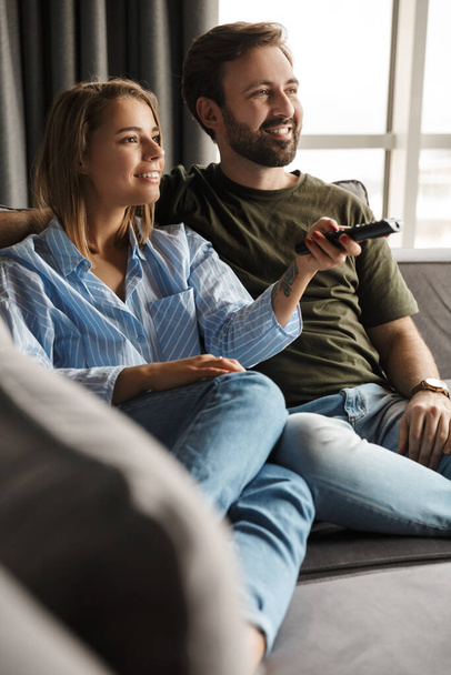Photo of joyful beautiful nice couple watching TV and using remote control while sitting on sofa at home - Фото, изображение