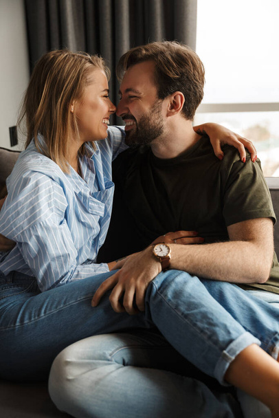 Photo of cheerful romantic caucasian couple smiling and hugging while sitting on sofa at home - Valokuva, kuva