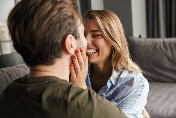Photo of cheerful romantic caucasian couple laughing and hugging while sitting on sofa at home - Foto, Bild