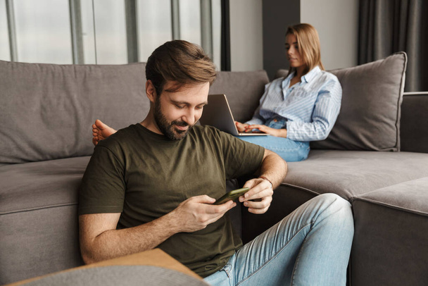 Photo of young caucasian focused couple using laptop and mobile phone while sitting at living room - Foto, afbeelding