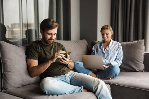 Photo of young caucasian joyful couple using laptop and mobile phone while sitting at living room - Valokuva, kuva