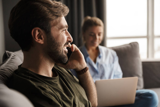 Foto de jovem casal caucasiano alegre usando laptop e telefone celular enquanto sentado na sala de estar
 - Foto, Imagem