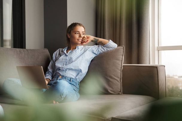 Image of beautiful young pleased woman using laptop and looking aside while sitting on sofa at living room - Fotoğraf, Görsel