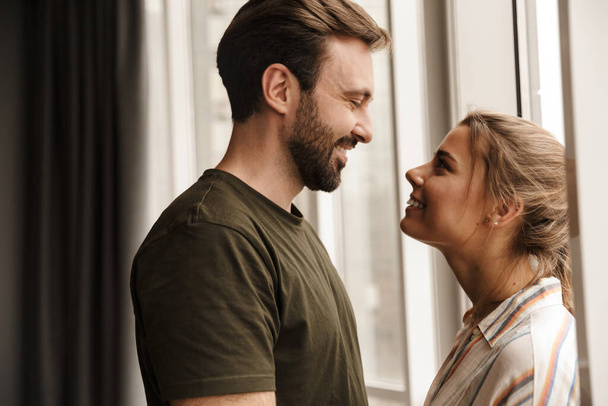 Photo of romantic caucasian cute couple smiling and looking at each other while standing near window at home - Fotó, kép