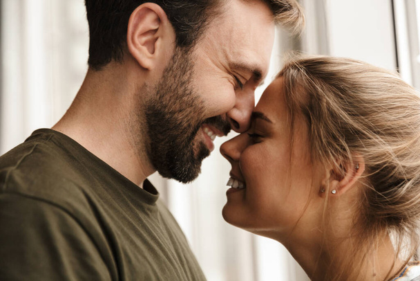Photo of romantic caucasian cute couple smiling and hugging while standing near window at home - Foto, Imagen