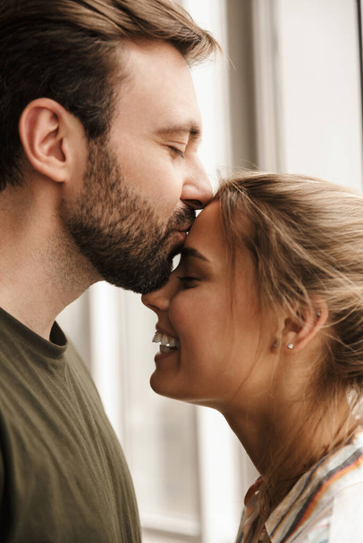 Foto de pareja linda romántica caucásica sonriendo y besándose mientras está de pie cerca de la ventana en casa
 - Foto, Imagen