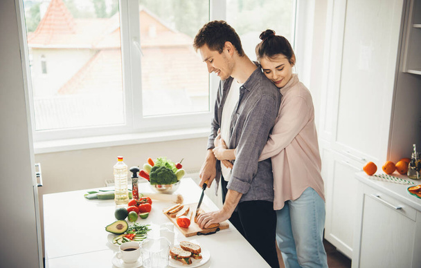 Brunette girl embracing her lover in the kitchen while making sandwiches - Photo, Image
