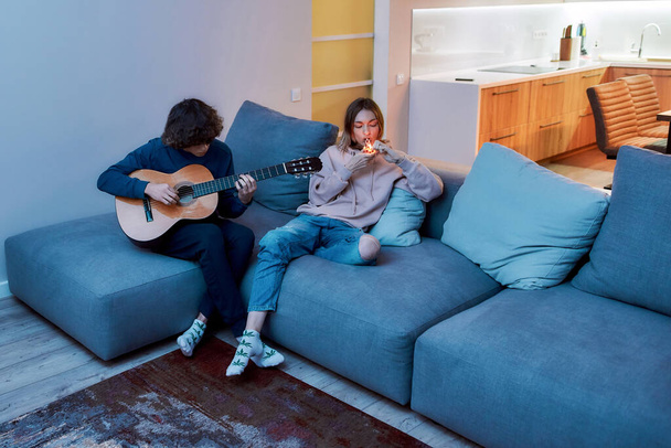 Friends relaxing. Young girl lighting cannabis in glass pipe or bong, resting with her boyfriend on sofa at home. Guy playing on the guitare - 写真・画像