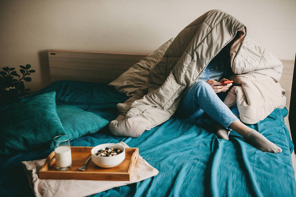 Caucasian woman covered with a quilt is chatting on phone before eating milk with cereals in bed - Foto, immagini