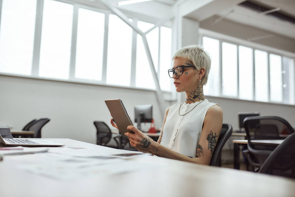 Using digital tools. Young and beautiful tattooed business woman wearing glasses working with digital tablet while sitting at her workplace in the office - Fotografie, Obrázek