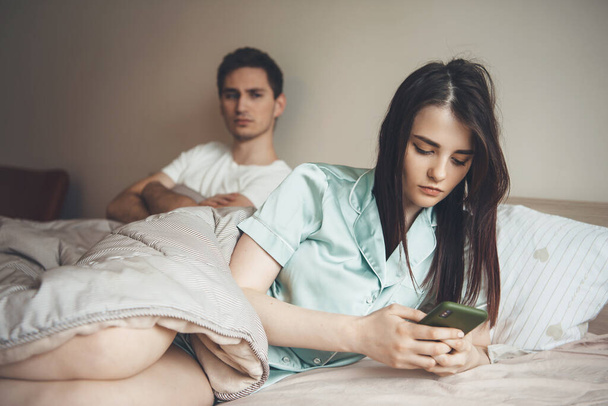 zaniepokojony biały mężczyzna patrząc na jego dziewczyna czatuje w łóżku na telefon komórkowy - Zdjęcie, obraz