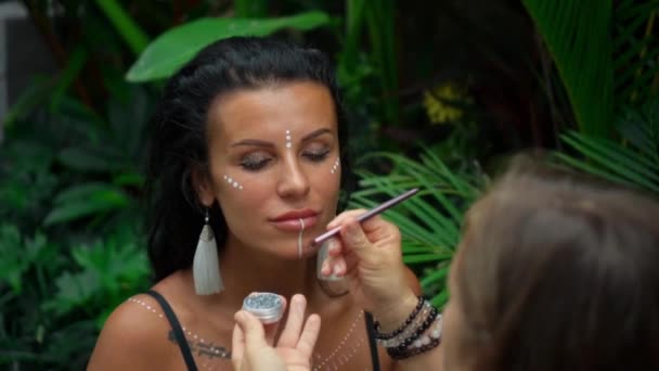 Video of a beautiful young brunette European girl with makeup and bodyart on the body sitting in a tropical green garden - Materiał filmowy, wideo