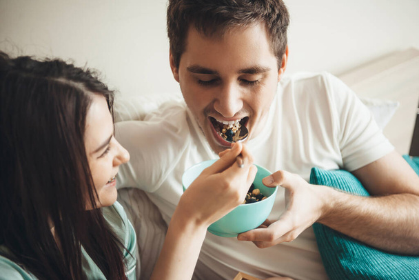 Χαριτωμένο καυκάσιο ζευγάρι τρώει δημητριακά στο κρεβάτι νωρίς το πρωί αγκαλιάζοντας και χαμογελώντας μαζί - Φωτογραφία, εικόνα