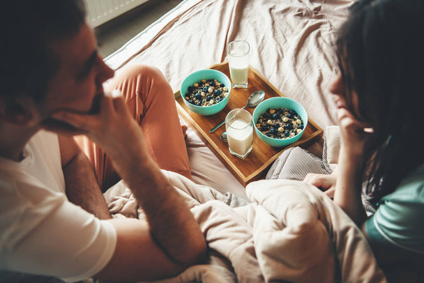 Υπέροχο ζευγάρι στο κρεβάτι κοιτάζοντας ο ένας τον άλλο πριν από την κατανάλωση δημητριακών με γάλα το πρωί - Φωτογραφία, εικόνα