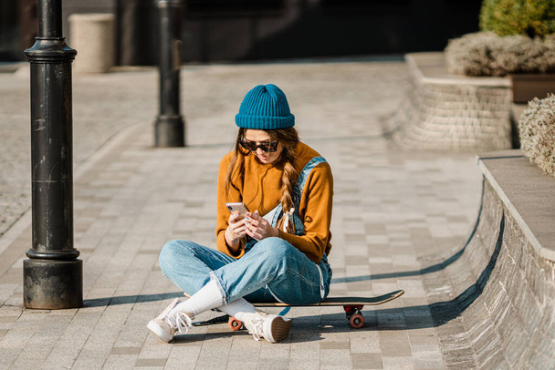 Young stylish caucasian woman skateboarder in a hat, sunglasses and jeans is using a phone next to a skate board in the city. Theme of active youth recreation in an urban environment. - Foto, imagen