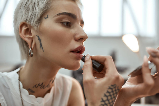 Looking perfect. Portrait of young and stylish blonde tattooed business woman applying lipstick while sitting in the modern coworking space - Fotó, kép