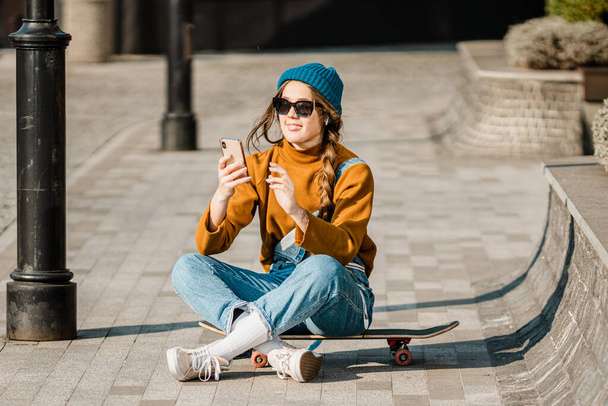 Young stylish caucasian woman skateboarder in a hat, sunglasses and jeans is using a phone next to a skate board in the city. Theme of active youth recreation in an urban environment. - Fotografie, Obrázek