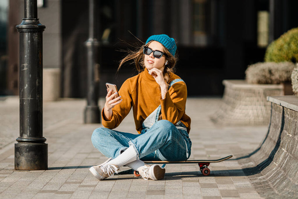 Fiatal, elegáns, kaukázusi nő gördeszkás kalapban, napszemüvegben és farmerben telefonál egy gördeszka mellett a városban. Az aktív ifjúsági kikapcsolódás témája városi környezetben. - Fotó, kép