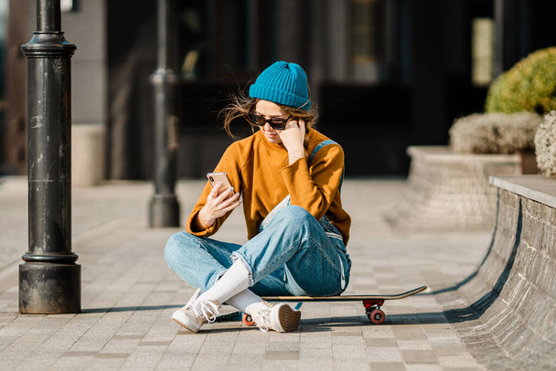 Girl sitting on skateboard and use mobile phone. Outdoors, urban lifestyle. cute skater girl sitting on skate board checking smart phone listening to music using internet and takes a photo. - Photo, image