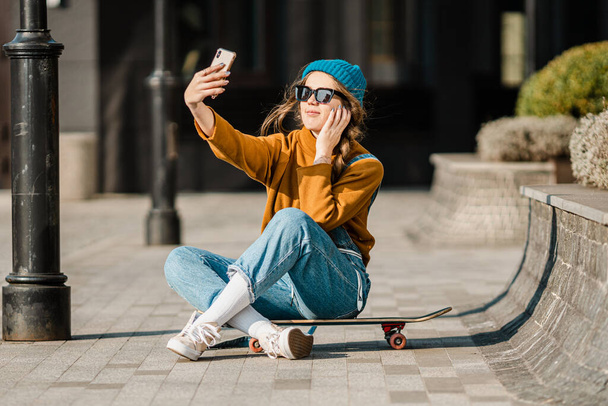 Cute urban girl outdoors with skateboard using smart phone. Happy young woman with skateboard, headphones and cell phone. Hipster female sit on skate board and using mobile phone. - Foto, imagen