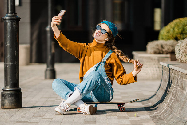 Młoda kobieta siedząca na deskorolce, korzystająca z telefonu. Kobieta na deskorolce w mieście. kobieta skate boarder ze smartfonem i deskorolką. cute skater siedzi na pokładzie, sprawdzanie smartfona i korzystania z Internetu. - Zdjęcie, obraz
