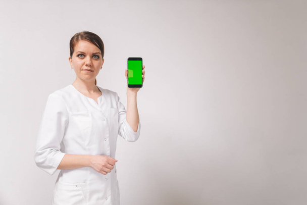 Beautiful young female doctor in a white coat on a light background holds a smartphone with a green screen. Isolated background for mobile application. - Foto, Imagem