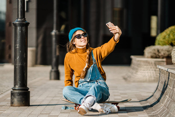 Dziewczyna siedzi na deskorolce i używa telefonu komórkowego. Na zewnątrz, miejski styl życia. ładny łyżwiarz dziewczyna siedzi na deskorolce sprawdzając smartfon słuchając muzyki za pomocą Internetu i robi zdjęcie. - Zdjęcie, obraz