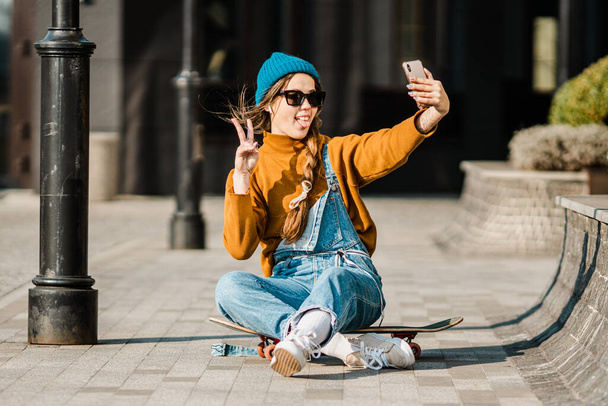 Young woman sitting on skateboard, using phone. skateboarding woman in city. female skate boarder with smartphone and skateboard. cute skater sitting on board, checking smart phone and use internet. - Photo, Image